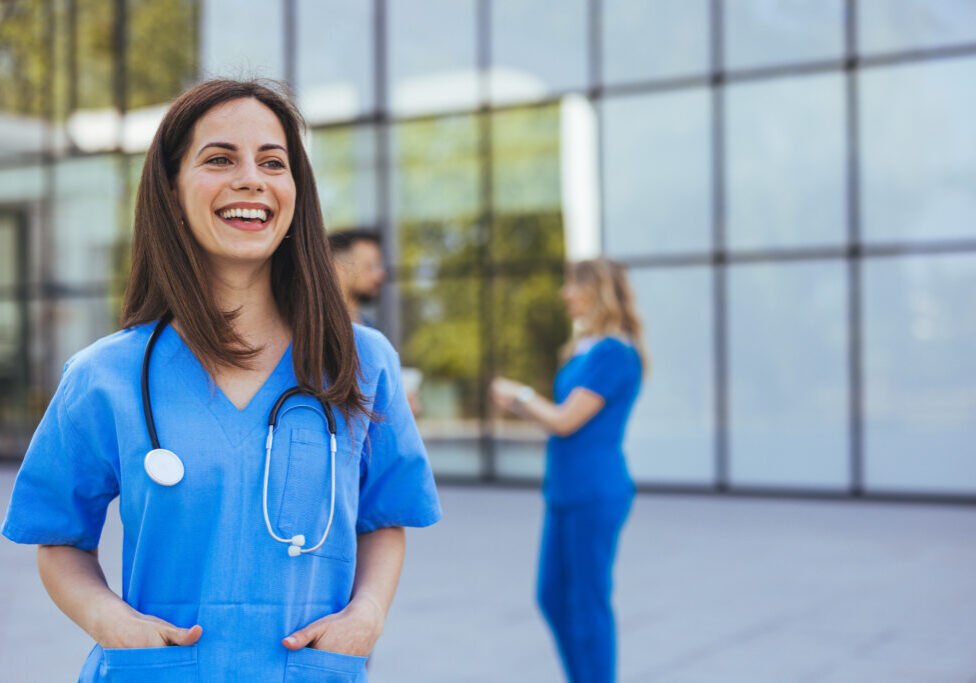 A joyful Caucasian nurse in blue scrubs stands confidently outside a hospital, stethoscope around her neck, with her colleagues in the background.